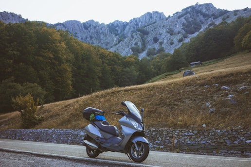 A motorcycle is parked on the side of a rural road surrounded by mountainous terrain. The road is bordered by a stone barrier, and beyond it, there is a grassy area leading up to dense forests and rocky hills. The scene is serene, with natural beauty highlighting the rugged landscape.