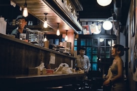 A dimly lit bar with a warm and cozy atmosphere. Several people are present, engaged in conversation. The lighting consists of hanging bulbs that create a moody ambiance. The shelves behind the counter are stocked with various bottles, and there are paper decorations hanging in the background.