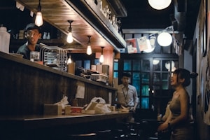 A dimly lit bar with a warm and cozy atmosphere. Several people are present, engaged in conversation. The lighting consists of hanging bulbs that create a moody ambiance. The shelves behind the counter are stocked with various bottles, and there are paper decorations hanging in the background.