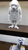 A white monkey sculpture holding a sign that reads 'DO NOTHING YOU'LL LIVE LONGER' is placed on a wooden shelf. Below it, a separate sculpture of a hand holding a bunch of white roses is positioned on a stack of books.