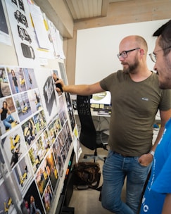Two individuals are engaging in a discussion in an office setting. They are examining a board filled with various printed images, some of which feature a yellow scooter. The office has a modern design with wooden elements and an organized workspace. Both individuals appear focused and are likely discussing the content on the board.