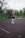 A person stands on an outdoor tennis court holding a tennis racket high in their right hand. They are wearing a black sweater, light gray pants, and white sneakers. The surrounding environment includes a fence, some trees, and a building in the background, suggesting a park or public recreational area. The scene appears overcast with fallen leaves scattered on the court.
