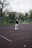 A person stands on an outdoor tennis court holding a tennis racket high in their right hand. They are wearing a black sweater, light gray pants, and white sneakers. The surrounding environment includes a fence, some trees, and a building in the background, suggesting a park or public recreational area. The scene appears overcast with fallen leaves scattered on the court.