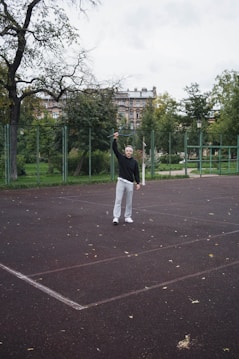 A person stands on an outdoor tennis court holding a tennis racket high in their right hand. They are wearing a black sweater, light gray pants, and white sneakers. The surrounding environment includes a fence, some trees, and a building in the background, suggesting a park or public recreational area. The scene appears overcast with fallen leaves scattered on the court.
