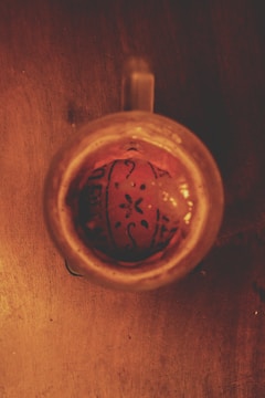 A brown ceramic mug is viewed from above on a wooden surface. Inside the mug is an intricate design resembling a flower or pattern with dark lines on a reddish-brown background.