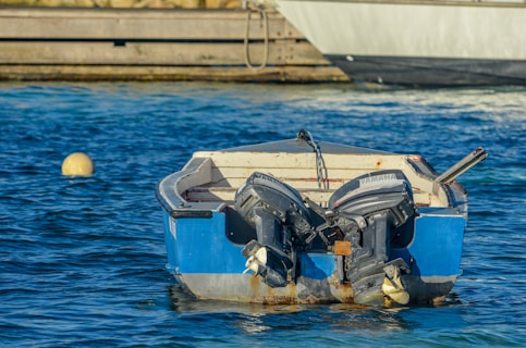 A small blue boat with two outboard motors is floating on clear blue water. The boat is secured with a chain, and a yellow buoy is visible nearby. In the background, part of a larger vessel and a wooden dock can be seen.
