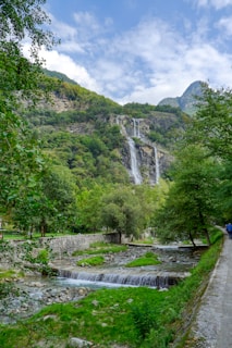A picturesque waterfall cascades down a lush, green mountainside, surrounded by trees and foliage. A serene stream flows through the foreground, with small stone walls lining a pathway. The sky is partly cloudy, adding depth to the peaceful landscape.