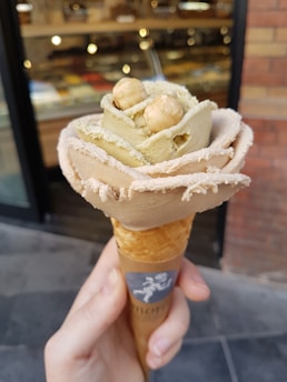A close-up of a hand holding an ice cream cone shaped like a flower, with two hazelnuts on top. The ice cream has a creamy texture and is artistically arranged in layers to resemble petals. The background is blurred, showing what appears to be a shop window.