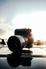 A professional camera with a large lens is placed on a reflective surface, with sunlight glowing behind it, creating a silhouette effect. The background features a bright sky with soft clouds.