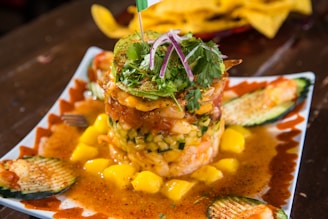 A vibrant dish featuring a tower of seafood and diced vegetables, garnished with fresh herbs and avocado slices. Surrounding the base are artistic swirls of sauce and sliced cucumbers, with scattered chunks of mango adding a pop of color.