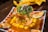 A vibrant dish featuring a tower of seafood and diced vegetables, garnished with fresh herbs and avocado slices. Surrounding the base are artistic swirls of sauce and sliced cucumbers, with scattered chunks of mango adding a pop of color.