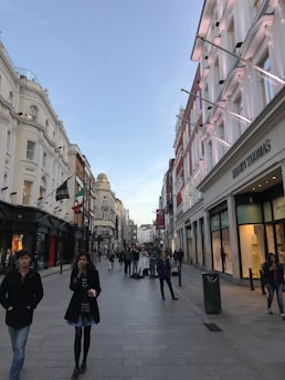 A bustling urban street scene with people walking along a pedestrianized shopping area. The street is lined with elegant multi-story buildings featuring various retail stores, including one named Brown Thomas. The atmosphere is lively, with groups of people casually strolling and interacting. The sky is clear, suggesting a pleasant day.