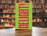 A vibrant novel cover featuring bold pink and black typography on a wooden shelf.
