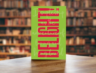 A book with a green cover featuring bold, vertical red text and cursive white writing overlaying the main title. The book is placed on a wooden table with a blurred background of bookshelves filled with books in a library setting.