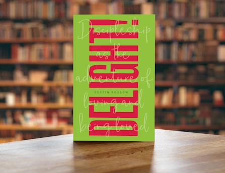 A book with a green cover featuring bold, vertical red text and cursive white writing overlaying the main title. The book is placed on a wooden table with a blurred background of bookshelves filled with books in a library setting.