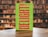 A book with a green cover featuring bold, vertical red text and cursive white writing overlaying the main title. The book is placed on a wooden table with a blurred background of bookshelves filled with books in a library setting.