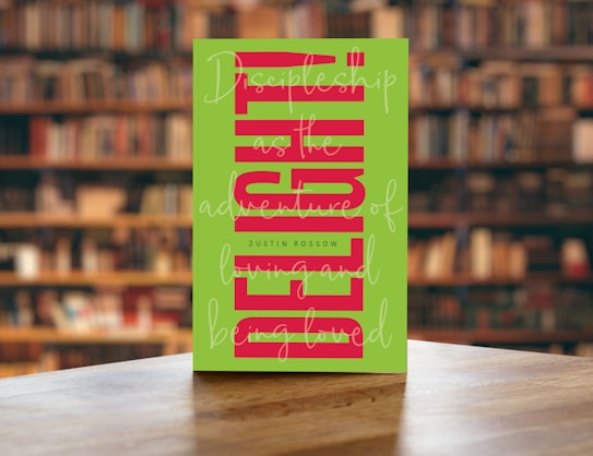 A book with a green cover featuring bold, vertical red text and cursive white writing overlaying the main title. The book is placed on a wooden table with a blurred background of bookshelves filled with books in a library setting.