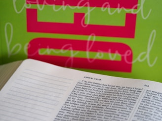An open book displaying a passage from John 15:9 is placed in the foreground, while a bright green background features bold pink text. The text overlays white script that reads 'loving and being loved.'