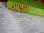 An open Bible lies on a surface, with the pages in focus. Resting on top is a bright green book titled 'DELIGHT!' in bold pink letters. The book cover includes cursive white text that reads 'Discipleship as a Way of Loving and...' The composition highlights religious and inspirational themes.