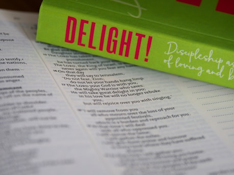 An open Bible lies on a surface, with the pages in focus. Resting on top is a bright green book titled 'DELIGHT!' in bold pink letters. The book cover includes cursive white text that reads 'Discipleship as a Way of Loving and...' The composition highlights religious and inspirational themes.