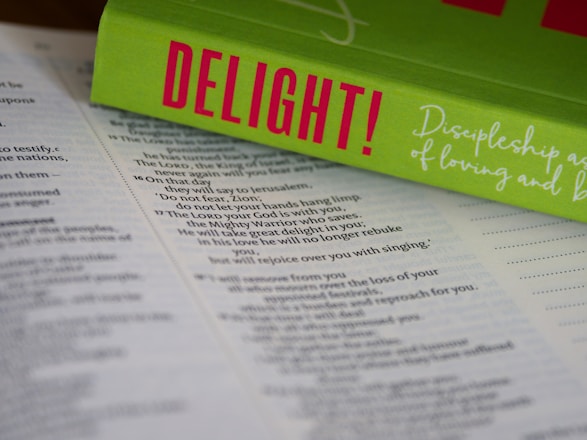 An open Bible lies on a surface, with the pages in focus. Resting on top is a bright green book titled 'DELIGHT!' in bold pink letters. The book cover includes cursive white text that reads 'Discipleship as a Way of Loving and...' The composition highlights religious and inspirational themes.