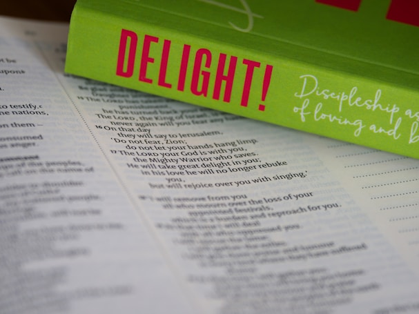 An open Bible lies on a surface, with the pages in focus. Resting on top is a bright green book titled 'DELIGHT!' in bold pink letters. The book cover includes cursive white text that reads 'Discipleship as a Way of Loving and...' The composition highlights religious and inspirational themes.