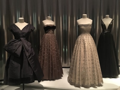 Four elegant evening gowns are displayed on mannequins, each with unique designs and silhouettes. The attire varies in color and style, featuring intricate detailing such as embroidery and delicate fabrics. The scene is set against a sleek, metallic background that adds a modern touch.
