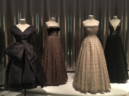 Four elegant evening gowns are displayed on mannequins, each with unique designs and silhouettes. The attire varies in color and style, featuring intricate detailing such as embroidery and delicate fabrics. The scene is set against a sleek, metallic background that adds a modern touch.