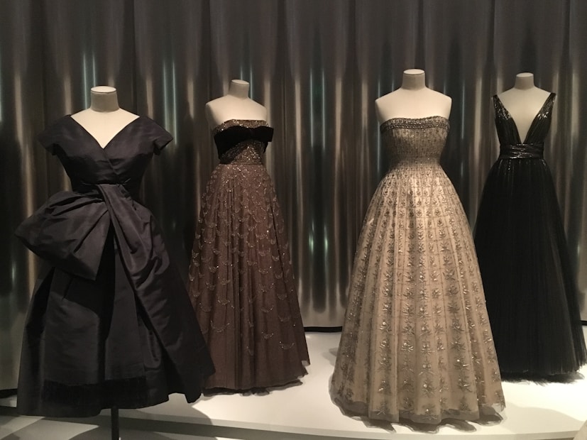 Four elegant evening gowns are displayed on mannequins, each with unique designs and silhouettes. The attire varies in color and style, featuring intricate detailing such as embroidery and delicate fabrics. The scene is set against a sleek, metallic background that adds a modern touch.