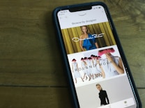 A smartphone displaying a fashion browsing interface. The screen shows images of models wearing designer clothing, with names like Oscar de la Renta and Roland Mouret prominently displayed. The phone is placed on a wooden table.