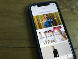 A smartphone displaying a fashion browsing interface. The screen shows images of models wearing designer clothing, with names like Oscar de la Renta and Roland Mouret prominently displayed. The phone is placed on a wooden table.