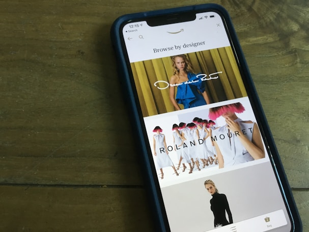 A smartphone displaying a fashion browsing interface. The screen shows images of models wearing designer clothing, with names like Oscar de la Renta and Roland Mouret prominently displayed. The phone is placed on a wooden table.