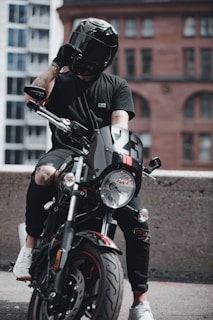A person wearing a black helmet and black clothing is seated on a black motorcycle. The motorcycle has the number 23 on its front and is positioned in an urban setting with a mix of modern and old-style buildings in the background. The person has tattoos on their right arm and is adjusting their helmet with their left hand.