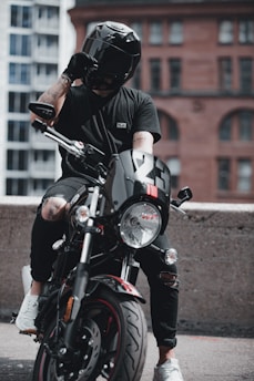 A person wearing a black helmet and black clothing is seated on a black motorcycle. The motorcycle has the number 23 on its front and is positioned in an urban setting with a mix of modern and old-style buildings in the background. The person has tattoos on their right arm and is adjusting their helmet with their left hand.