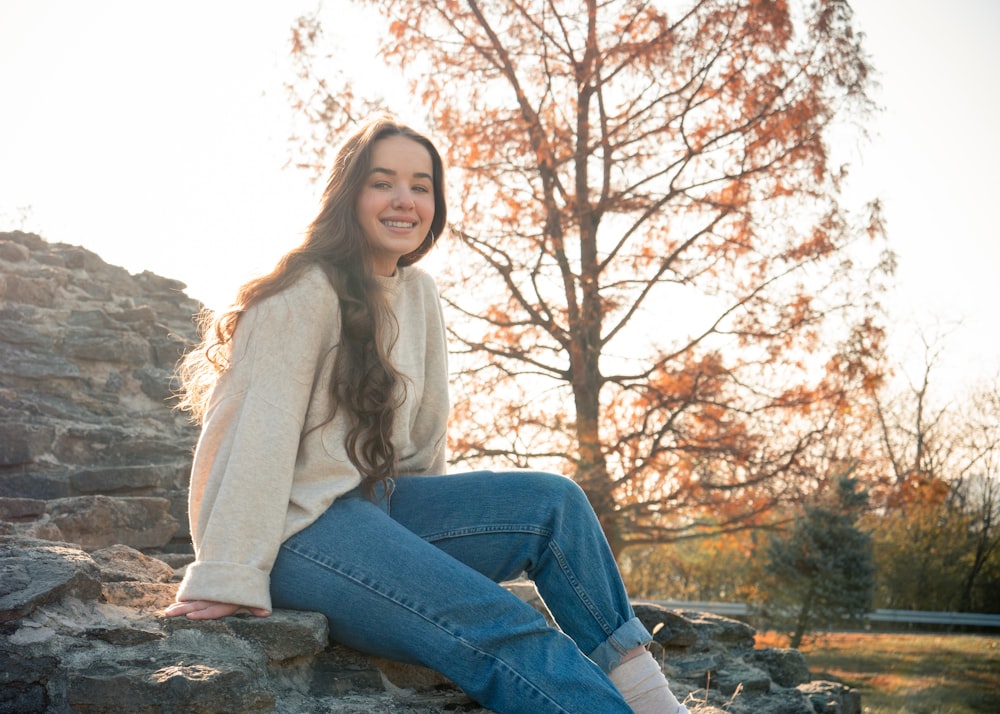 Frau in weißem Langarmhemd und blauer Jeans sitzt auf Felsen