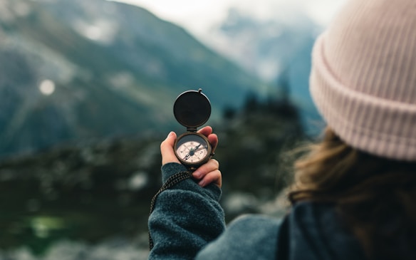 A person is holding a compass in their hand, pointing it towards a blurred scenic background of mountains. The individual is wearing a warm, green jacket and a light-colored beanie, suggesting a cool atmosphere.