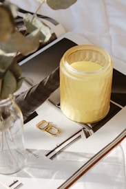 An amber-colored candle in a textured glass jar sits on top of an open magazine. To the left, there is a glass vase containing eucalyptus leaves. A pair of golden hoop earrings is placed near the candle.