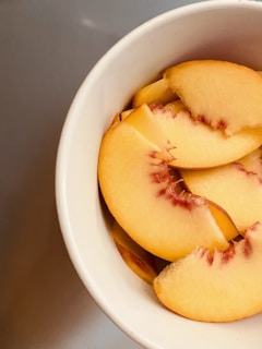 Slices of peach neatly arranged in a white bowl. The fruit has a soft, velvety skin with shades of orange and pink, and the fleshy interior is light orange with red patches near the pit.