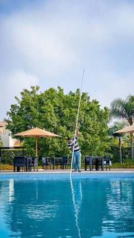A person cleaning a swimming pool using a long pole, surrounded by patio furniture and large umbrellas on a sunny day. In the background, lush green trees and a blue sky are visible, creating a serene outdoor atmosphere.