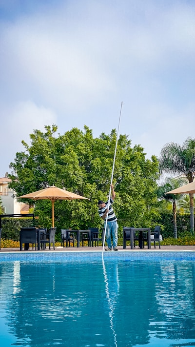 A person cleaning a swimming pool using a long pole, surrounded by patio furniture and large umbrellas on a sunny day. In the background, lush green trees and a blue sky are visible, creating a serene outdoor atmosphere.