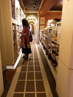 A person wearing a colorful traditional outfit stands in a narrow, ornate bookstore aisle. The shelves are filled with numerous books on both sides. The ceiling is decorated with intricate designs, and the lighting creates a warm ambiance. Decorative arches and floral motifs add to the aesthetic.
