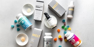 An arrangement of skincare products, supplements, and gummies on a light, textured surface. The products include bottles, boxes, and plates with samples. Some gummy candies are scattered around, adding color.