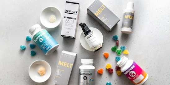 An arrangement of skincare products, supplements, and gummies on a light, textured surface. The products include bottles, boxes, and plates with samples. Some gummy candies are scattered around, adding color.