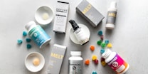 An arrangement of skincare products, supplements, and gummies on a light, textured surface. The products include bottles, boxes, and plates with samples. Some gummy candies are scattered around, adding color.