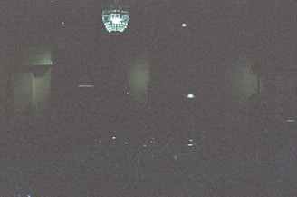 A dimly lit room with visible noise and grain in the image, featuring a chandelier hanging from the ceiling. The room is mostly dark, with only a few light sources illuminating parts of the ceiling.
