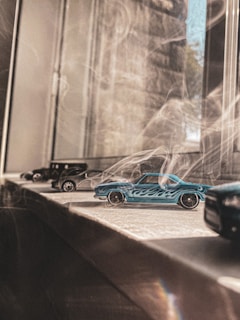 A collection of small toy cars, including a prominent blue one with flame designs, is positioned on a windowsill. Sunlight filters through the window, casting soft shadows and adding a warm tone to the scene.