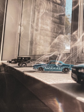 A collection of small toy cars, including a prominent blue one with flame designs, is positioned on a windowsill. Sunlight filters through the window, casting soft shadows and adding a warm tone to the scene.