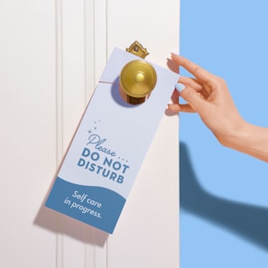 A hand with neatly manicured nails is placing a 'Please Do Not Disturb' sign on a door handle. The sign emphasizes 'Self care in progress' and is decorated with small stars. The environment is serene and minimalistic with a soothing color palette.