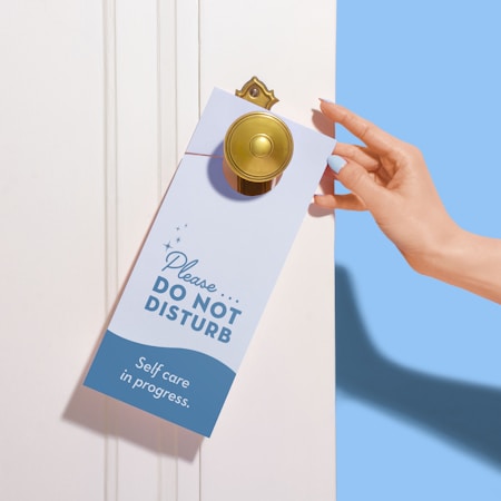 A hand with neatly manicured nails is placing a 'Please Do Not Disturb' sign on a door handle. The sign emphasizes 'Self care in progress' and is decorated with small stars. The environment is serene and minimalistic with a soothing color palette.
