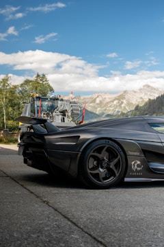 A sleek, high-performance sports car with a matte black finish is parked on a paved surface. The car has distinctive alloy wheels and aerodynamic features. In the background, there is a large truck with industrial equipment, set against a backdrop of lush green trees and towering mountains under a clear blue sky.